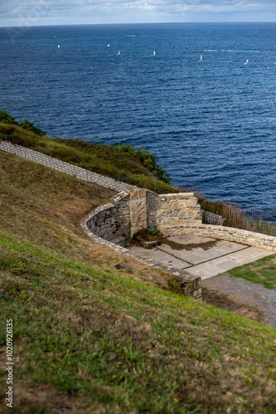 Obraz A picturesque coastal scene featuring a stone amphitheater perched on a cliff overlooking a vast ocean. The amphitheater is surrounded by lush green grass and stone walls, providing a stunning panoram