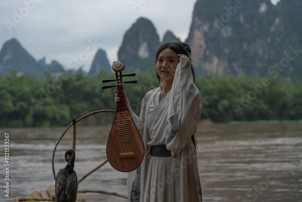 Obraz Hanfu girl poses with Pipa on bamboo raft with a mountainous background. Xingping, China