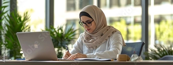 Fototapeta A Muslim woman in a hijab works at her desk with a laptop, writing notes on paper, working online or studying for school