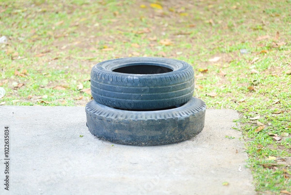 Obraz A close-up shot of two old car tires stacked on top of each other, placed on a paved surface outdoors. The background shows a grassy area with scattered dry leaves, indicating an outdoor setting.