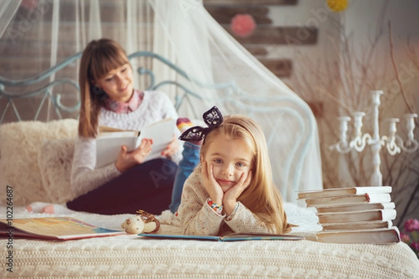 Obraz Portrait of a smiling young cute mother and daughter reading a book lying and relax on the floor in a bright big white room next to the window