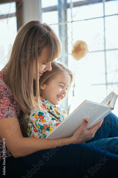 Fototapeta Portrait of a smiling young cute mother and daughter reading a book lying and relax on the bed in a bright big white room 
