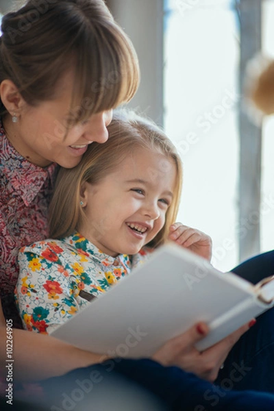 Obraz Portrait of a smiling young cute mother and daughter reading a book lying and relax on the bed in a bright big white room 