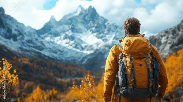 Fototapeta A man wearing a yellow jacket and a backpack is standing in front of a mountain