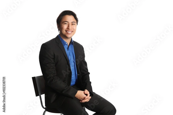 Fototapeta Man in his 20s sitting down on a chair against a white background, wearing a black suit with a dark blue shirt. Looking at camera with a big toothy smile.