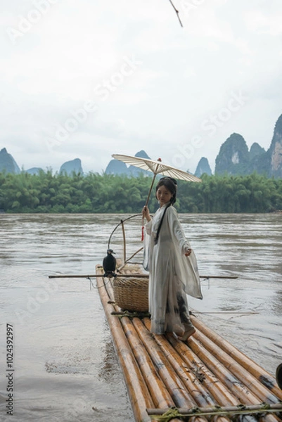 Obraz Woman in Hanfu attire holds an umbrella while standing on a bamboo raft in Xingping, China
