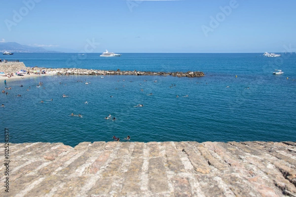 Fototapeta Looking over the ancient walls of Antibes in the south of France to the beach, yachts and people swimming in the sea