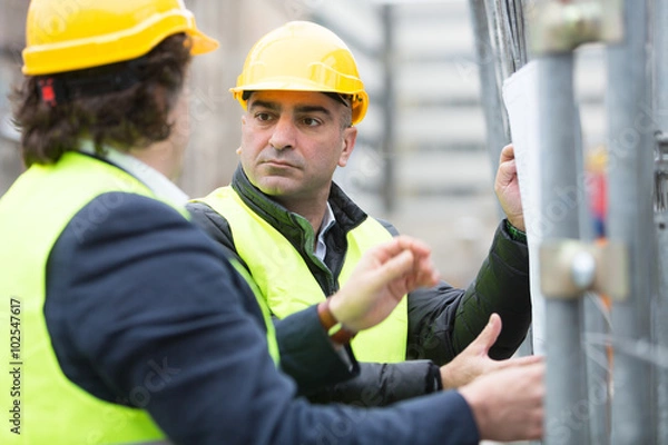Fototapeta Two architects wearing hardhat and yellow jacket reading office blueprints against scaffolding at construction site