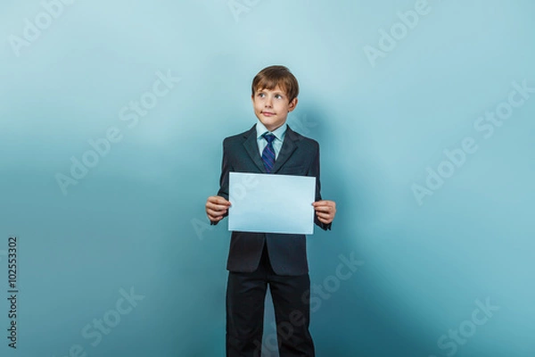 Obraz a boy of twelve  European appearance in a suit  holding a blank sheet on a gray  background