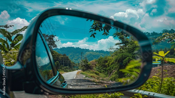 Fototapeta A rearview mirror reflects a scenic drive with lush greenery, mountains, and a bright sky, creating a picturesque travel moment.
