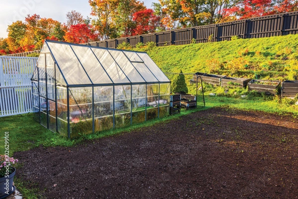 Fototapeta Greenhouse prepared for winter with stored items, set in an autumn garden with colorful trees and empty flowerbeds. Sweden.