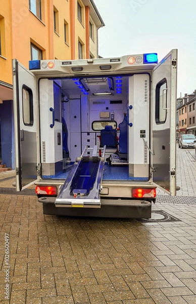 Fototapeta An ambulance with its rear door open and a patient stretcher extended stands outside a residential building in Germany, waiting for a patient. Vertical format.