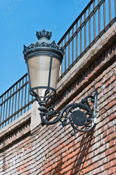 Fototapeta An antique street lamp with a decorative wrought iron bracket and a glass globe. The lamp is mounted on a brick wall and is partially shaded by the shadows of the iron railings above.