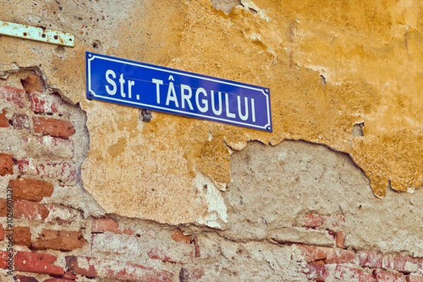 Fototapeta A close-up of a blue street sign for "Str. Târgului" mounted on a weathered brick wall. The wall is covered in peeling paint and has visible cracks and damage. 