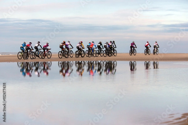 Fototapeta unrecognizable people drive with mountain bikes on a beach during a contest. with a visible reflection in the water in the foreground. 