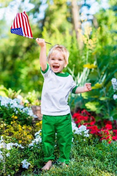 Obraz Barefoot little boy with blond hair laughing and waving american flag in sunny park or garden on summer day. Portrait of child on blurred background. Independence Day, Flag Day concept