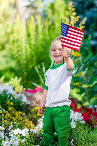 Obraz Smiling blond little boy holding american flag and waving it in sunny park or garden on summer day. Portrait of child on blurred background. Independence Day, Flag Day concept