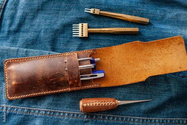 Fototapeta rustic-style leather pencil case containing a few pens, laid open on a blue denim surface alongside additional leathercraft tools, evoking a sense of handcrafted durability and timeless craftsmanship
