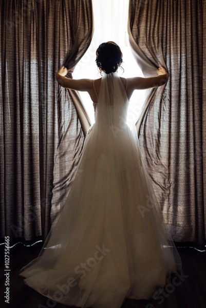 Fototapeta The girl stands with her back near the window and opens the curtains with her hands