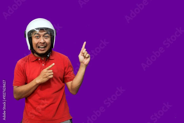Fototapeta Young Asian driver man wearing helmet looking up while raising both hands and pointing upwards with happy expression
