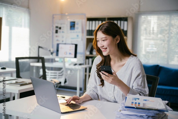 Fototapeta Smiling businesswoman is working on a laptop and using a smartphone at her desk in a modern office. She is surrounded by paperwork and digital devices