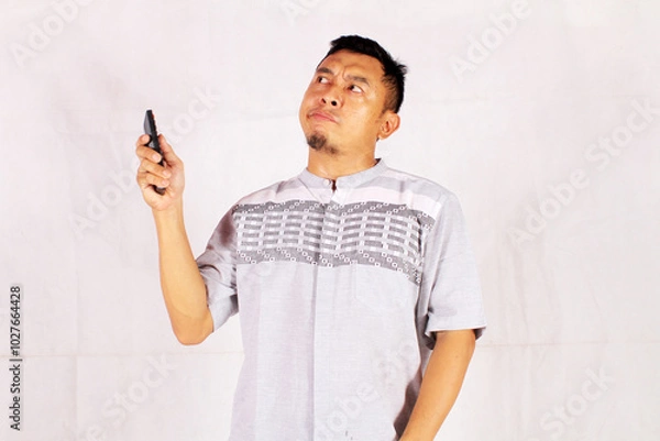 Fototapeta Asian man holding tv remote with surprised and confused expression
