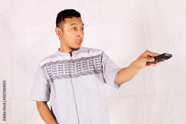 Fototapeta Asian man holding tv remote with surprised and confused expression
