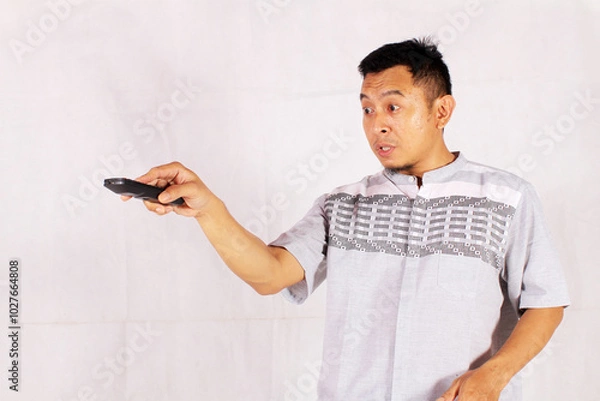 Fototapeta Asian man holding tv remote with surprised and confused expression
