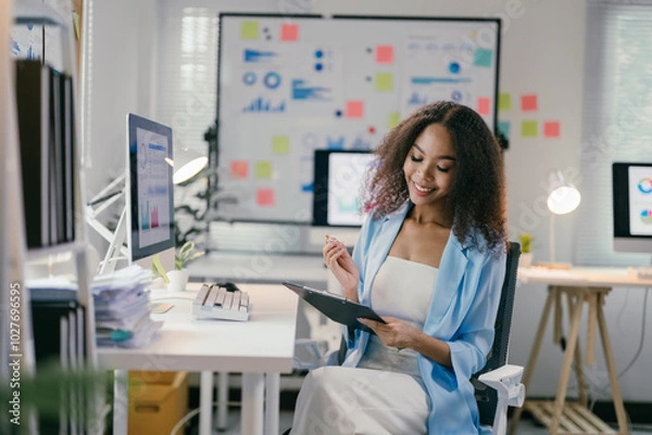 Fototapeta African american businesswoman is sitting at her desk reviewing documents with a thoughtful expression. With a bright smile, she appears to be content with her work
