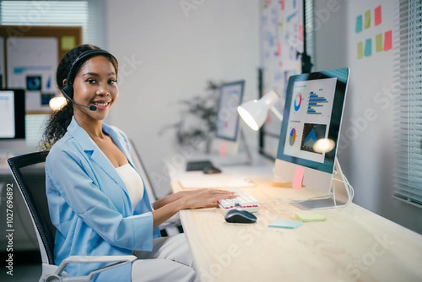 Fototapeta Confident african american businesswoman working joyfully at sleek desk in bustling office, fully engaged with computer, showcasing professionalism and expertise in corporate world