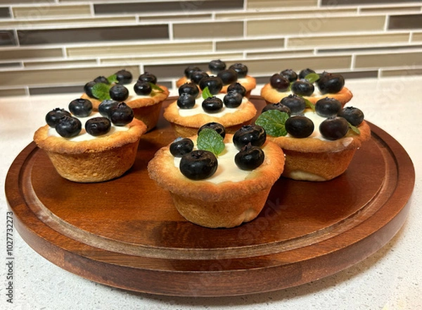 Obraz A batch of freshly made blueberry tartlets sit beautifully on a wooden tray. Each tartlet has a golden-brown crust, filled with smooth vanilla custard, and is topped with plump blueberries.