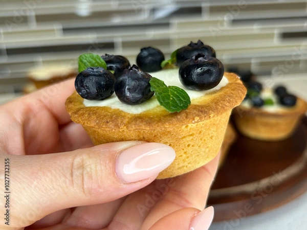 Obraz A delicate blueberry tartlet is held in hand, showcasing its crisp, buttery crust, creamy vanilla filling, and fresh blueberry topping. The pop of mint adds a refreshing twist to the look.