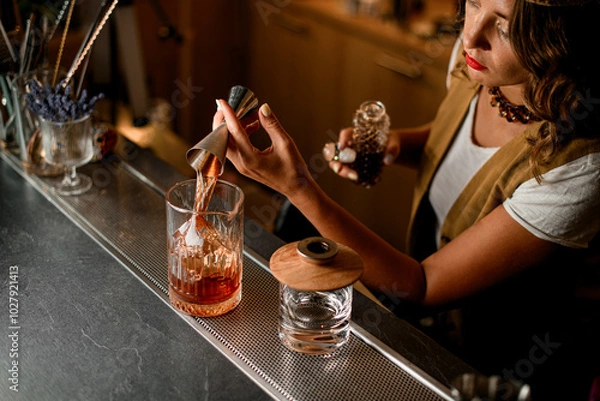 Fototapeta Top view of mixing glass in which bartender pours brown drink from jigger