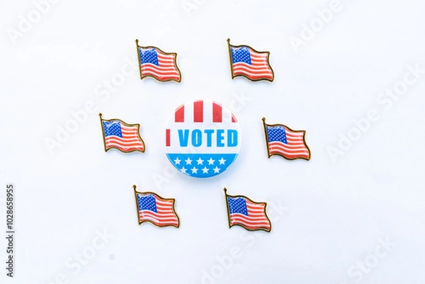 Fototapeta An "I Voted" button surrounded by six small American flag pins arranged in a circular pattern on a white background. The button features red, white, and blue colors with stars, representing the voting