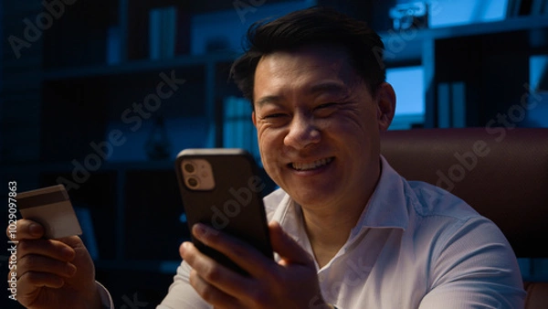 Fototapeta Smiling happy Asian man at dark night evening home office with cellphone using credit card Korean male businessman make payment booking order carefree with easy online mobile phone shopping discount