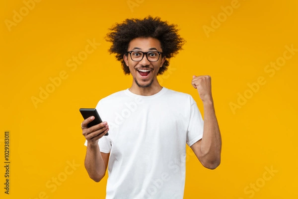 Fototapeta Studio portrait of young bearded African American man wearing basic white shirt holding in hand mobile cell phone and doing winners gesture with excited face expression celebrating clenching fist sayi