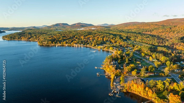 Obraz A stunning sunrise over Schroon Lake, NY, with warm light casting a soft glow on the calm water and surrounding autumn-colored forests and mountains.