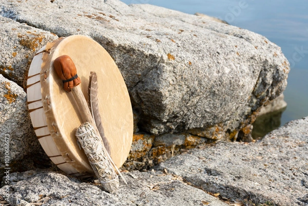 Fototapeta An image of a hand made leather meditation drum leaning on a rock face with sacred feather and sage smudge stick.