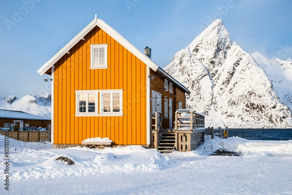 Fototapeta Traditional yellow rorbu house and Olstind mountain peak in Sakrisoy fishing village on Lofoten islands, Norway. Winter landscape with snowy mountains and Scandinavian cottage