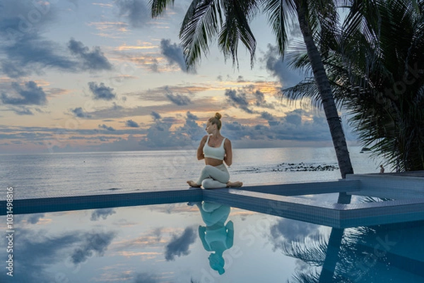 Fototapeta Young woman in a white clothes doing yoga on the beach near  swimming pool early morning in the summer. Sunrise sky in the background .