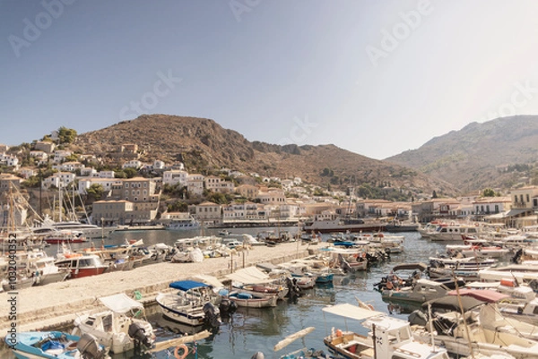 Obraz The vibrant harbor of Hydra Island showcases numerous boats docked in calm waters, surrounded by quaint buildings and steep hills under a clear blue sky on a sunlit day.