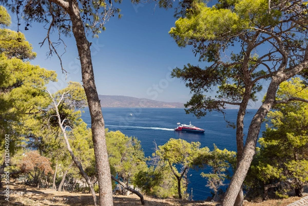 Obraz A colorful boat glides through the clear blue waters off the coast of Hydra, Greece, surrounded by vibrant green trees under a bright sunny sky.