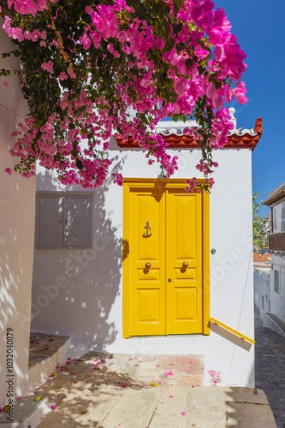 Obraz A striking yellow door stands out against the white walls, adorned with blooming bougainvillea, creating a picturesque setting in Hydra, Greece during the day.