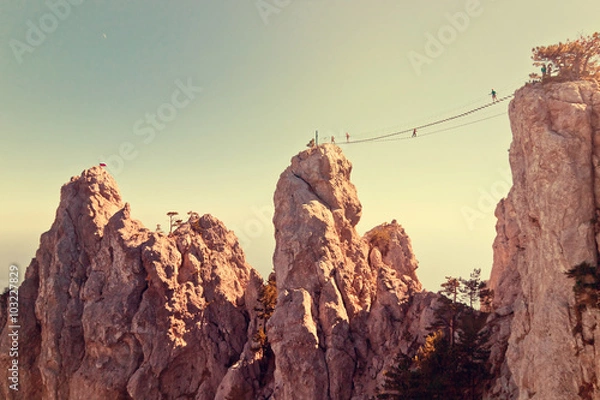 Fototapeta People crossing the chasm on the rope bridge. Black sea background, Crimea, Russia. Image with vintage filter