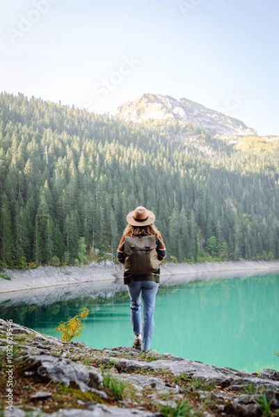 Obraz Time for solitude. Happy tourist woman enjoys the view of the mountain lake in sunny weather. Woman sitting on a rock in the mountains, resting on a halt on a hike.