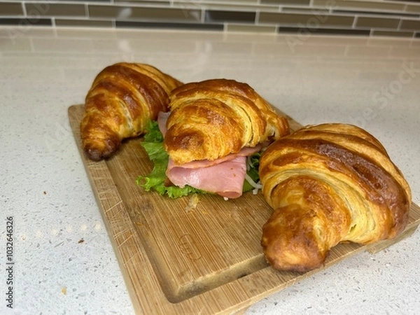 Obraz A delicious croissant sandwich filled with ham and lettuce, presented on a wooden cutting board in a modern kitchen setting. A medium-angle shot with a neutral background.