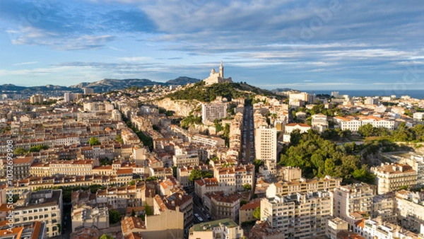 Fototapeta Aerial view of Marseille, France, featuring rooftops, streets, and the Notre-Dame de la Garde basilica, with the Mediterranean Sea in the background.