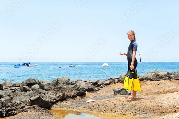 Fototapeta Young cheerful girl in diving suit with paddles standing on rock