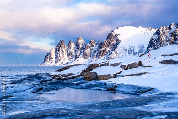 Fototapeta Tungeneset or Devils Jaw mountain on Senja island, Norway in winter. Winter landscape with snowy mountain peaks, fjord waters and ice coast in northern Norway. Scenic view with dramatic sky