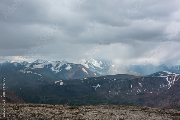 Fototapeta Dramatic top view from high stony pass to alpine valley against large mountain range with snow-capped peaks in gray rainy low clouds. Beautiful big snowy mountain tops in rain under grey cloudy sky.